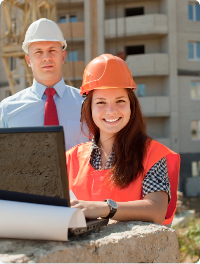Female construction worker in safety vest and hard hat stands next to a laptop with a male worker next to her.